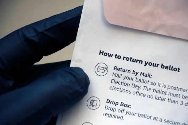 Closeup of vote-by-mail return ballot instructions on an official election mail envelope, being held in a person's hand wearing a black glove