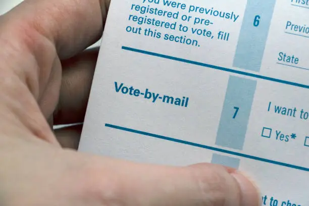 A hand holding a Voter Registration form, closeup on the 'Vote-by-Mail' section