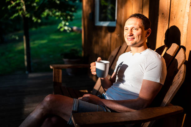 Man happy sitting relaxing on rocking chair lounge on porch of house in morning wooden cabin cottage drinking coffee or tea from cup mug Man happy sitting relaxing on rocking chair lounge on porch of house in morning wooden cabin cottage drinking coffee or tea from cup mug drinks on the deck stock pictures, royalty-free photos & images