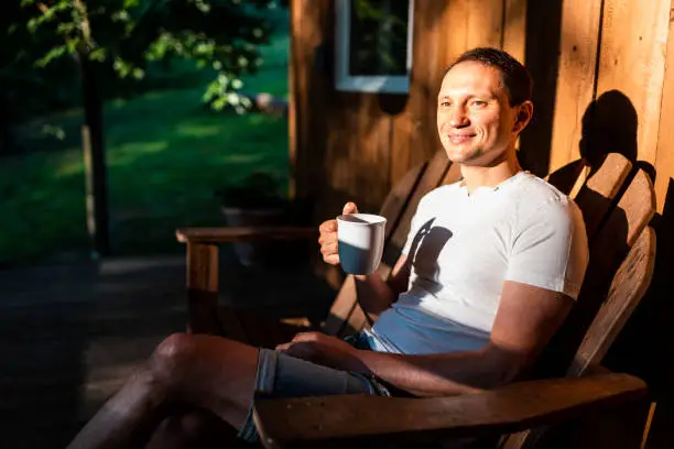 Photo of Man happy sitting relaxing on rocking chair lounge on porch of house in morning wooden cabin cottage drinking coffee or tea from cup mug