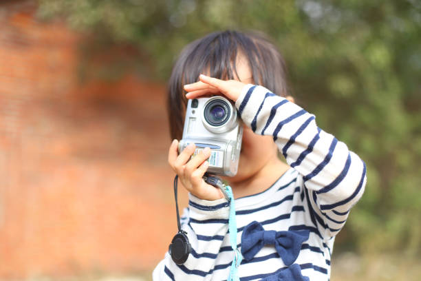 a smiling Asian girl take photos with a digital camera before a brick house.