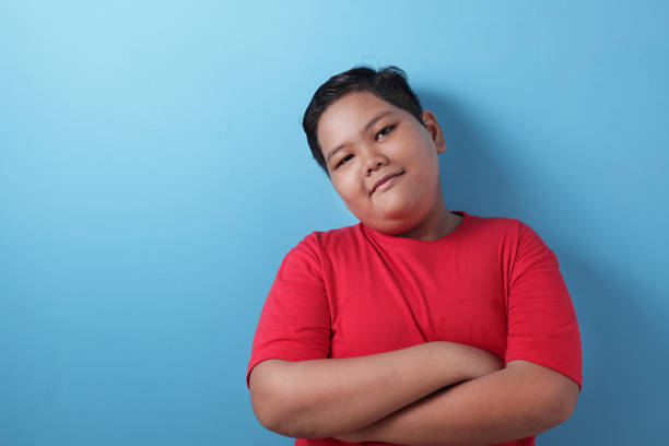 Fat Asian boy looks at the camera and smiles happily, crosses his arms in front of his chest Fat Asian boy looks at the camera and smiles happily, gestures confidently, crosses his arms in front of his chest, blue background with copy space overweight boy stock pictures, royalty-free photos & images
