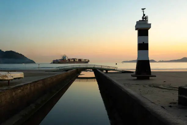 Photo of View of the city of Santos with channel 6 and its lighthouse with a cargo ship leaving the port during sunset
