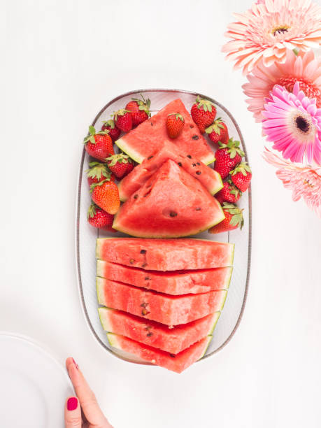 Watermelon slices with strawberries on ceramic tray on white wooden table with pink flowers. Fresh summer snack