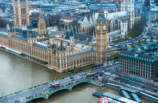 Aerial view of the city of London, overlooking multiple old buildings. In the middle the Big Ben clock tower. To the left the river of thames.