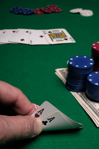 Close up vertical of a pair of aces held between thumb and forefinger on green felt. Poker chips and playing cards in background.