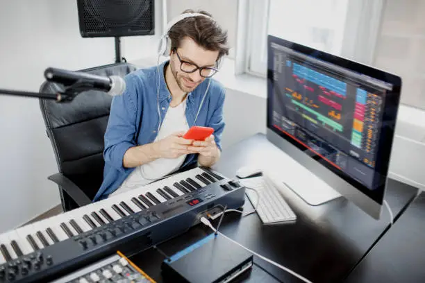 Photo of Man holds a smartphone in his hands and uses it. Male music arranger composing song on midi piano and audio equipment in digital recording studio. Man produce electronic soundtrack or track in project at home.