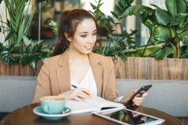 istock Young smiling asian girl student doing homework in notebook with tablet on table using mobile phone at cafe 1234736832