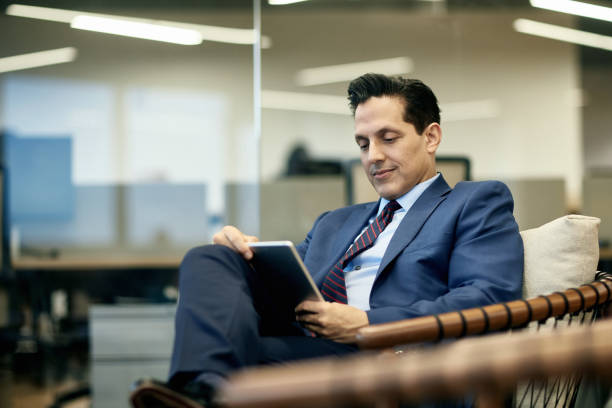 empresario hispano leyendo tableta digital en sillón - lobby businessman office building built structure fotografías e imágenes de stock