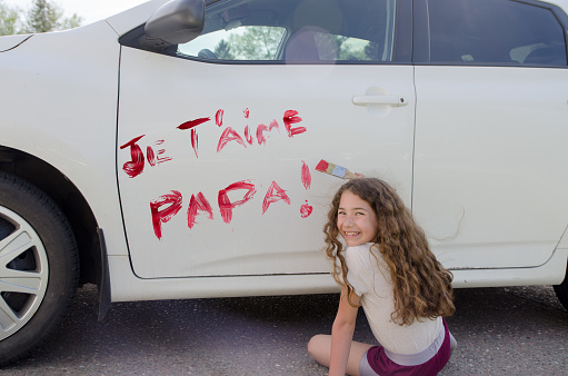 Little girl writing I love you dad on car door with red paint.
Text in French: Je t'aime papa