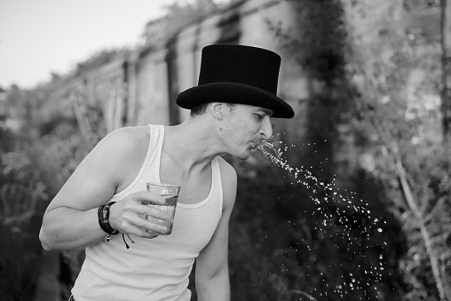 Three-quarter portrait of young man, wearing grey pants,white top, black classic hat, holding water glass, spitting on the side. Black and white picture of creative man on abandoned construction site.