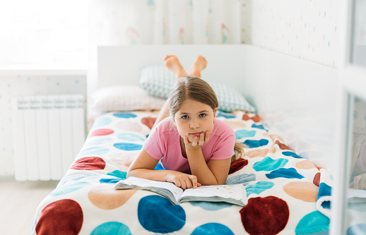 Cute tween girl in pink t-shirt reading book lying on bed in bright room at the home