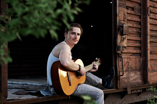 Young handsome man, wearing white t-shirt and grey pants, sitting inside old railway carriage, playing guitar, composing music Brunette male musician singer with guitar on abandoned train in summer.