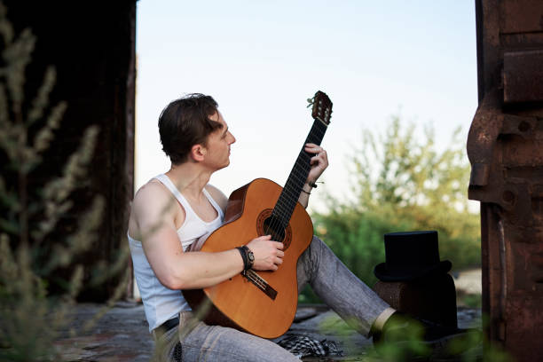 Young handsome man, wearing white t-shirt and grey pants, sitting inside old railway carriage, playing guitar, composing music Brunette male musician singer with guitar on abandoned train in summer.