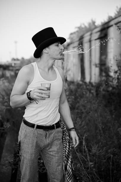 Three-quarter portrait of young man, wearing grey pants,white top, black classic hat, holding water glass, spitting on the side. Black and white picture of creative man on abandoned construction site.
