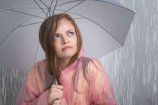 ピンクのレインコートを着た若い白人女性の女の子は、面白い��嫌な顔で大雨の下で傘を持っています。雨季コンセプト - monsoon ストックフォトと画像