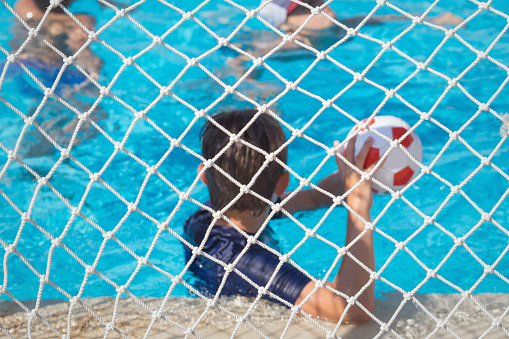 Children paly water polo with ball in swimming pool. View through goal net selective focus. Summer fun sports with copy space