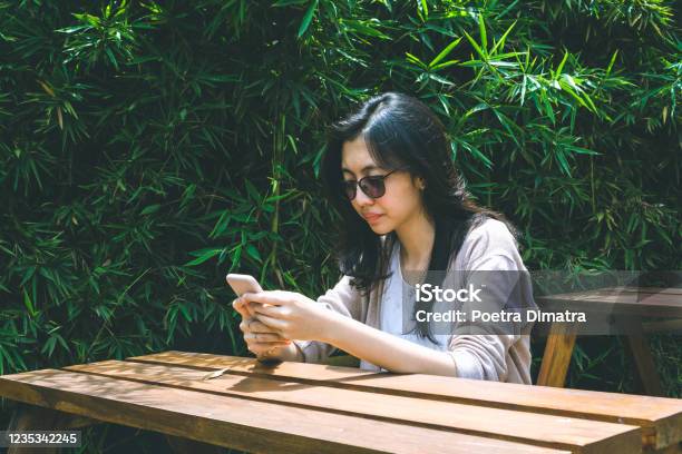 Asian Millennials Woman Hold Smartphone Sitting On Picnic Table Stock Photo - Download Image Now