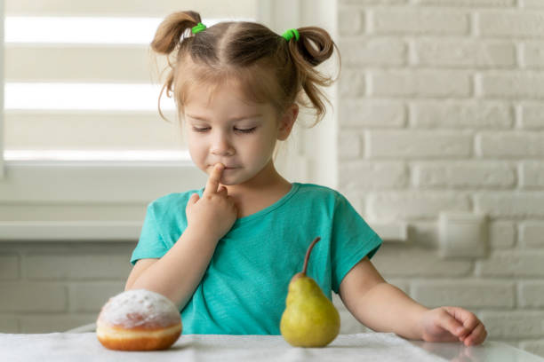 little girl chooses between donut and fruit. the concept of proper nutrition, the right choice - cake pick imagens e fotografias de stock