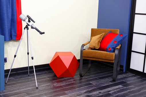 A brown chair with two pillows. Bright tones of the children's room, red, white, blue, brown. Children's toy, built-in duffcase cabinet. Telescope for the study of stars.