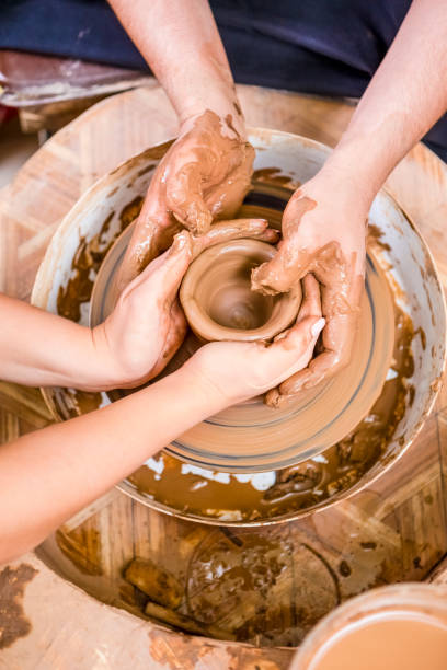 istock Professions Ideas. Old Hand Experienced Male Potter Working with Female Apprentice. Working with Clay Lump on Potter's Wheel in Workshop.Vertical Image 1235631511