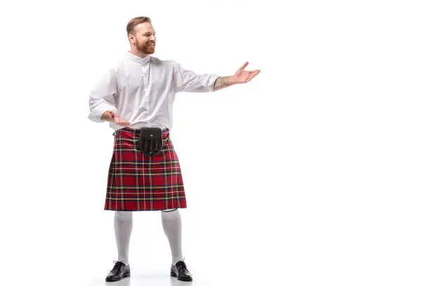 smiling Scottish redhead man in red kilt gesturing on white background