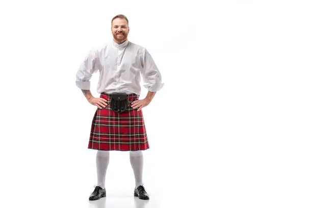 smiling Scottish redhead man in red kilt with hands on hips on white background