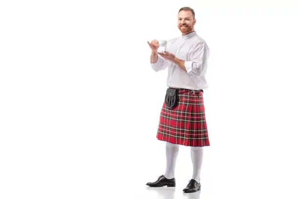 smiling Scottish redhead man in red kilt drinking coffee on white background