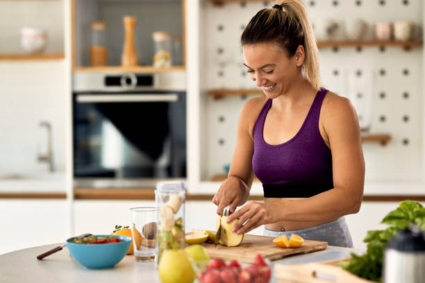 Happy athletic woman cutting fruit while preparing healthy meal in the kitchen. Young happy sportswoman slicing fruit while making smoothie in her kitchen. smoothie photos stock pictures, royalty-free photos & images