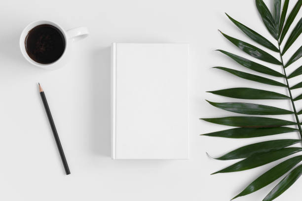 istock Top view of a white book mockup with  workspace accessories and a palm leaf on a white table. 1235866490
