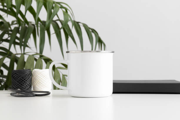 istock Enamel mug mockup with workspace accessories on a white table and a palm plant. 1235875144