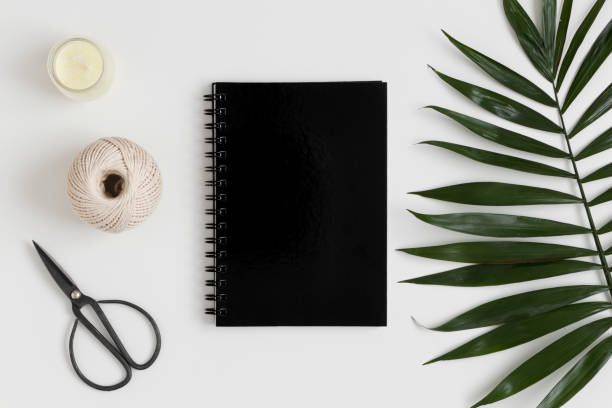 istock Top view of a black notebook mockup with  workspace accessories and a palm leaf on a white table. 1235878740