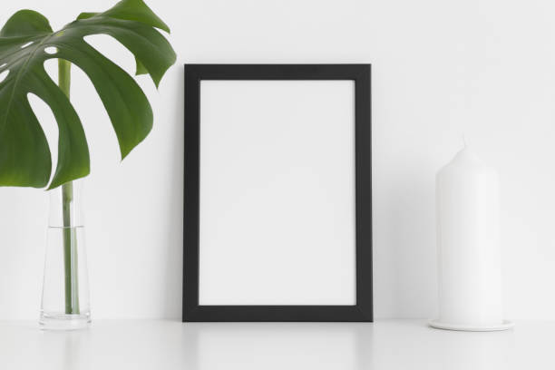 istock Black frame mockup with a candle and a monstera leaf in a vase on a white table.Portrait orientation. 1235903343