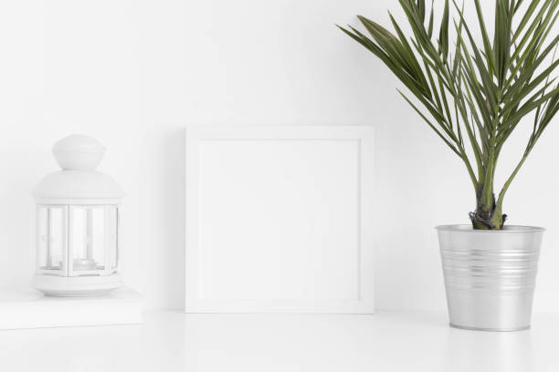 istock White square frame mockup with a palm in a pot, candle holder and a book on a white table. 1235904910