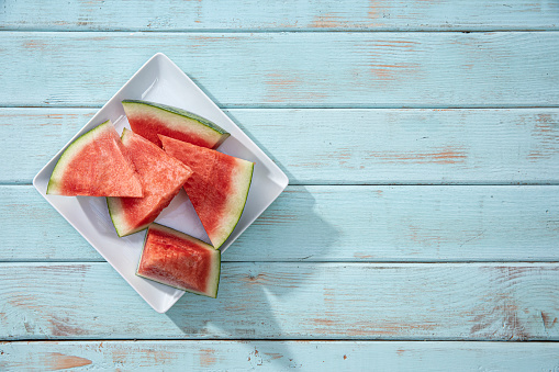 This is a photograph of triangle sliced watermelon on a white square plate sitting on a Blue retro wood picnic bench. This image would work well for the summer time and Fourth of July holiday