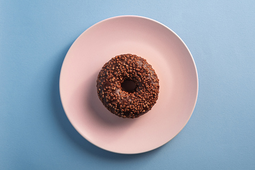 Chocolate donut with sprinkles on pink plate, sweet glazed dessert food on blue minimal background, top view