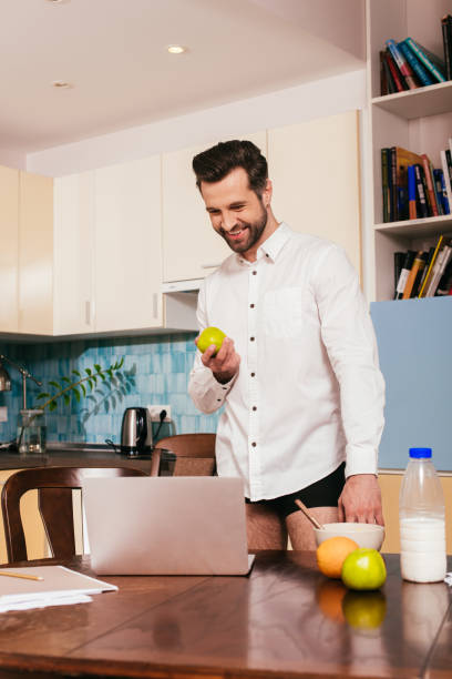 istock Selective focus of handsome man in panties and shirt smiling while holding apple near laptop on kitchen table 1236105607