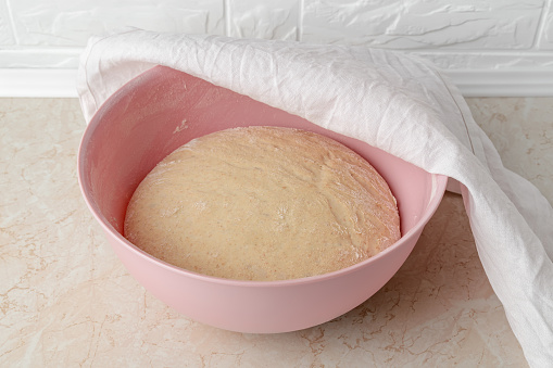 Fresh yeast dough rising in a large plastic bowl covered with white linen towel on the kitchen table. Hands preparing dough for baking homemade bread. Front view.