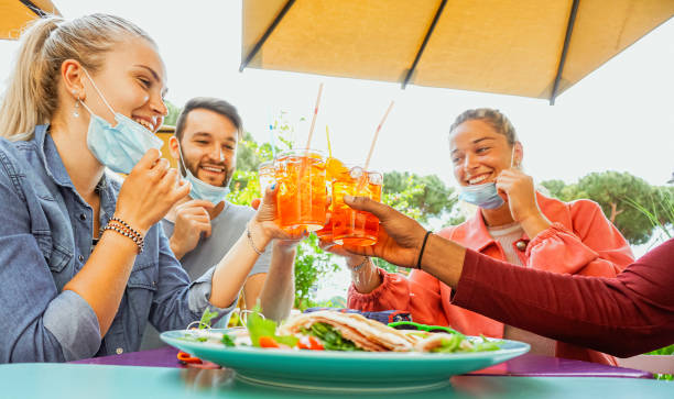 コロナウイルスから保護されるフェイスマスクを着用した夏の外のレストランバーでコックテールを飲んだり、スナックタパスを食べたりする友人たち - スプリッツで応援し、楽しんでいる� - restaurant sign sign language food ストックフォトと画像