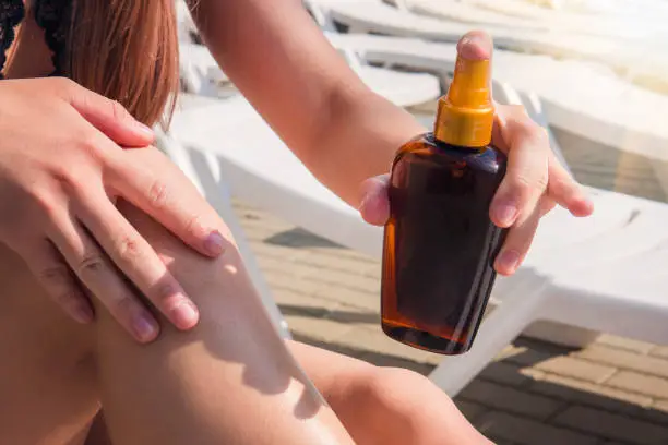 Lady is massaging sunscreen lotion while sunbathing. Female model during summer vacation. Sunscreen suntan lotion in spray bottle. Young woman in spraying tanning oil on her leg from bottle in hand.