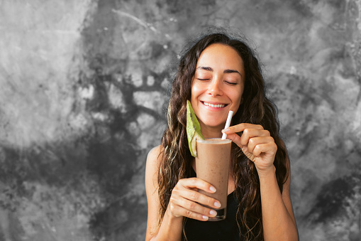 Funny happy woman drinking chocolate milk shake with cocoa and laughing. Summer drink concept. Gray concrete wall background.