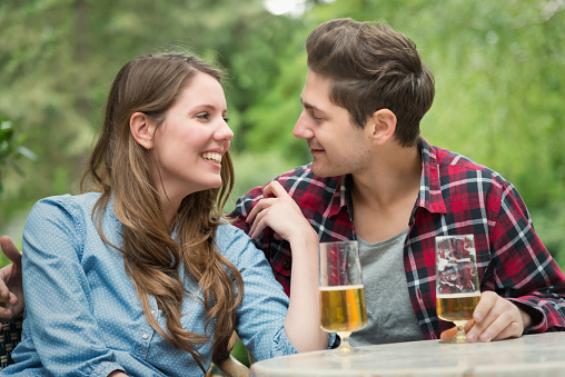 Young Couple sitting together side by side in Outdoor Beer Garden drinking a Beer, smiling at each other, talking, enjoying their time together. Young Couple Vacation Lifestlye Concept.