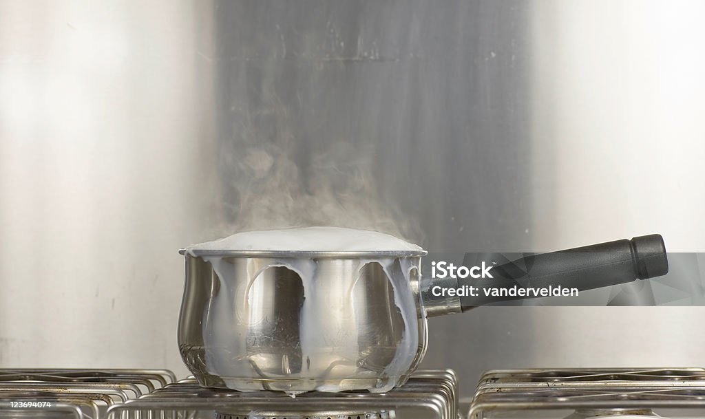 Saucepan Of Boiling Milk Steel and monochromatic study of milk boiling over on a gas hob. Overflowing Stock Photo