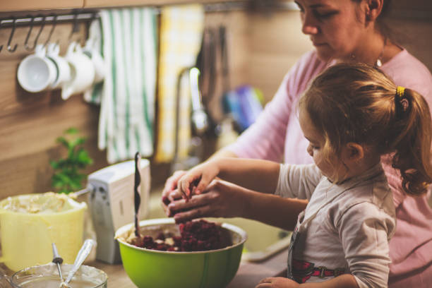 petite fille mélangeant des cerises dans la pâte à gâteau avec sa mère - two parent family indoors home interior domestic kitchen photos et images de collection