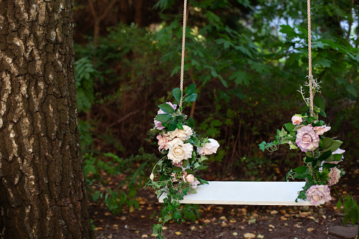 Wooden swing in playground outdoors. Empty swing placed in park. Wedding swing decorated with flowers roses. Garden swing hanging  from a large tree on green grass background. Garden decor. Romance