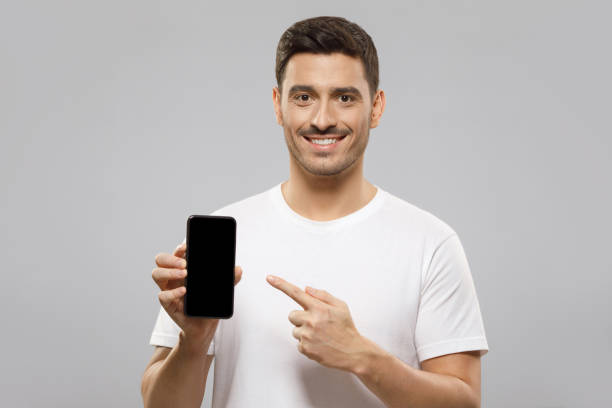 istock Young handsome man in white t-shirt holding smartphone, pointing to blank screen with finger, recommending mobile application, isolated on gray background with copy space 1237257807