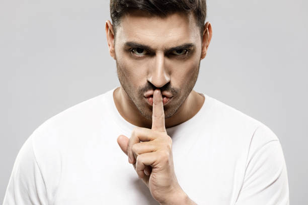 istock Young serious man in white t-shirt touching lips with finger, asking to stay silent and keep secret, showing shh gesture isolated on gray background 1237257811