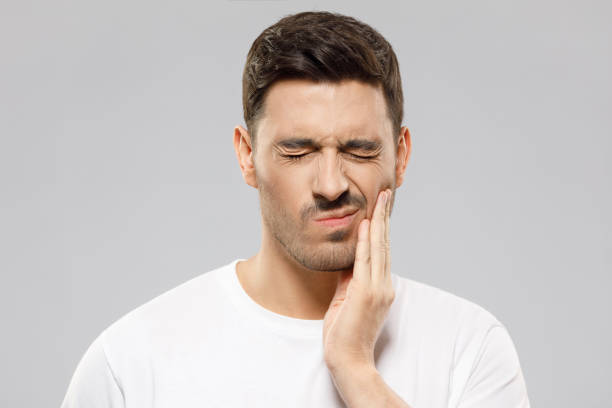 Young man in white t-shirt suffering from severe toothache, touching cheek with fingers, eyes closed because of strong pain, isolated on gray background Young man in white t-shirt suffering from severe toothache, touching cheek with fingers, eyes closed because of strong pain, isolated on gray background jaw pain stock pictures, royalty-free photos & images