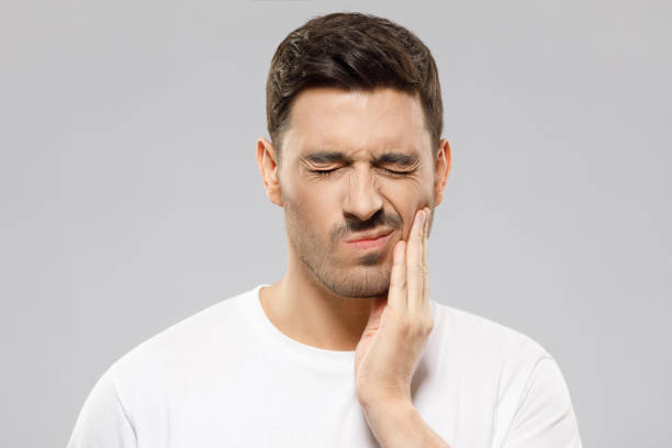 jovem de camiseta branca sofrendo de dor de dente grave, bochecha tocando com os dedos, olhos fechados por causa de dor forte, isolado em fundo cinza - queixo - fotografias e filmes do acervo