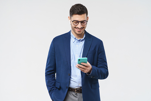 Young man dressed in blue blazer and shirt, wearing glasses, looking attentively at smartphone screen, smiling positively, isolated on gray background